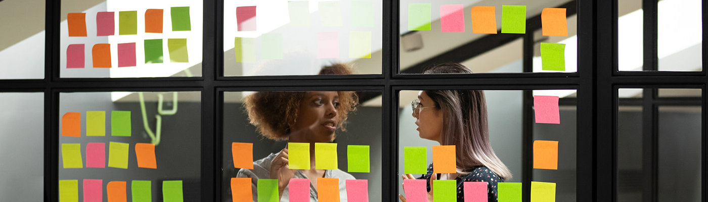 Header Image: Two female colleagues discuss their scrum board that is covered in post it notes