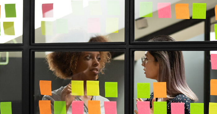 Header Image: Two female colleagues discuss their scrum board that is covered in post it notes