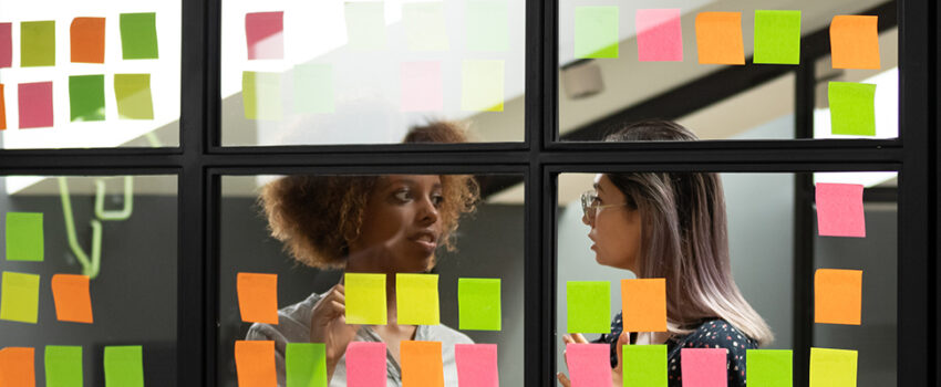 Header Image: Two female colleagues discuss their scrum board that is covered in post it notes