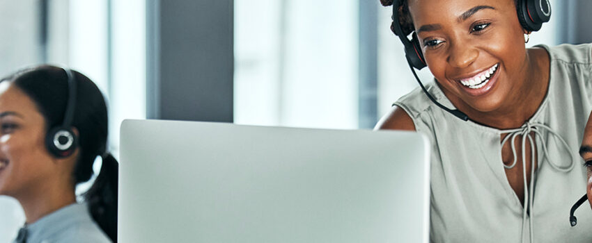 Header image of a contact center, where a woman wearing a headset is helping another man in a headset with his call.