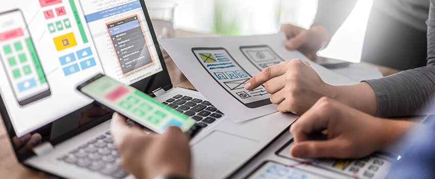 Two people lean over a laptop comparing a drawn mock up of a mobile app to the digital mock up, to the actual mobile experience on a cellphone