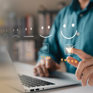 A man in a teal shirt sits in front of a laptop and chooses from a customer satisfaction rating that hovers above it.