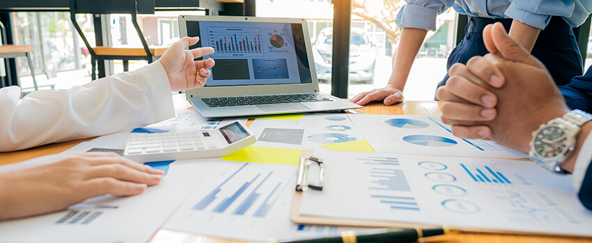 A table is strewn with reports showing pie charts and other data. A hand points at a laptop where more reporting is displayed.