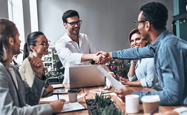 A diverse group of account executives sit around a table. One leans across to shake hands with a new client.