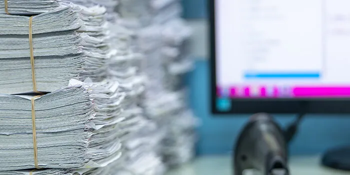 A massive pile of documents waiting to be scanned sit in front of a computer monitor.