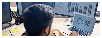 A man works on a laptop. Behind the laptop sits a monitor showing a bar chart.