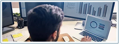 A man works on a laptop. Behind the laptop sits a monitor showing a bar chart.