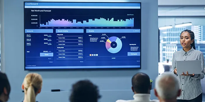 A woman gives a presentation to her colleagues while a report is displayed on the monitor behind her.