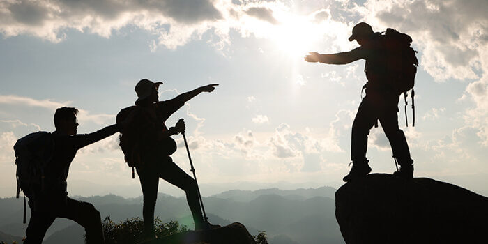 A silhouetted hiker atop a precipice helps his companions cross the gap.