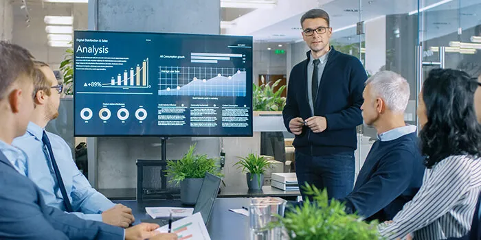 A young white man presents data on a large TV to a board meeting.