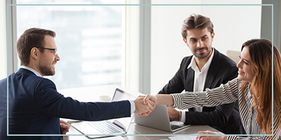 A woman reaches across the table to shake hands with a male sales person as another business man watches