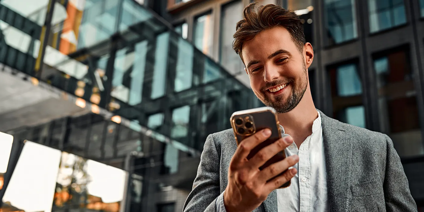 A young white businessman standing outside an office building smiles down at his phone