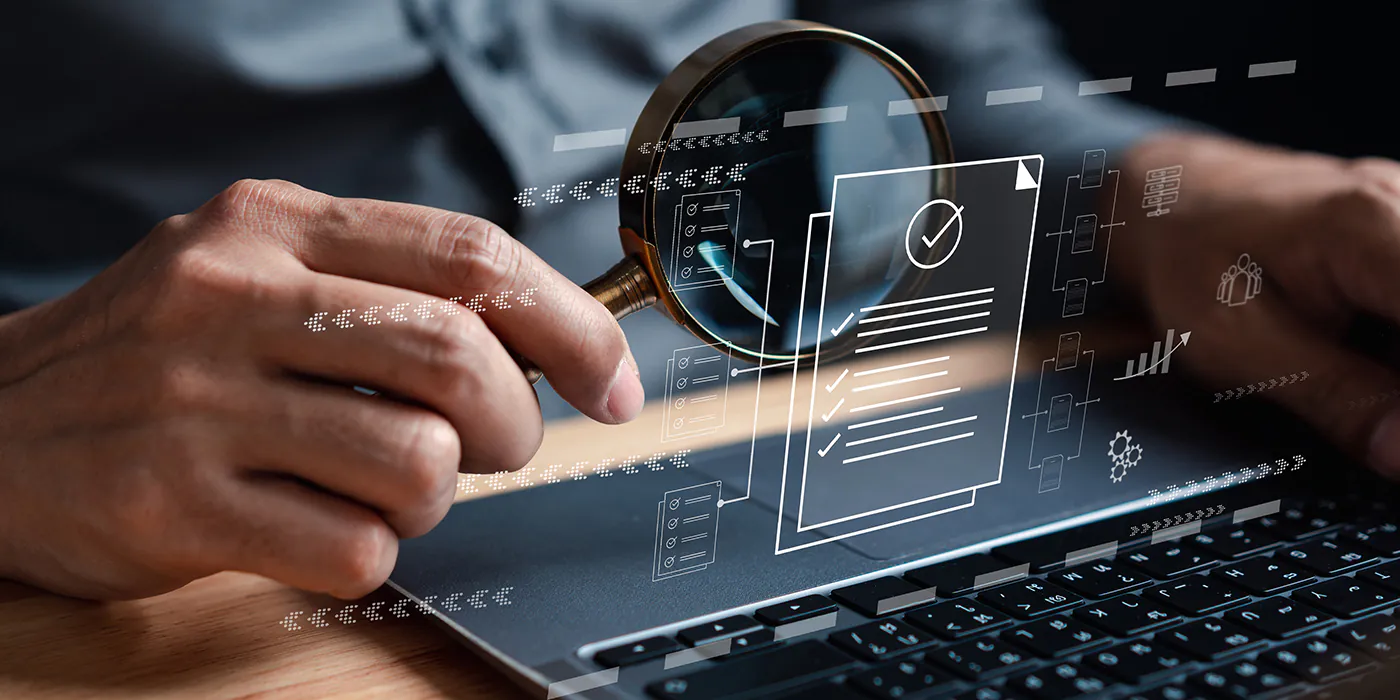 Hand holding magnifying glass over digital document icons on a laptop keyboard indicating data analysis and review